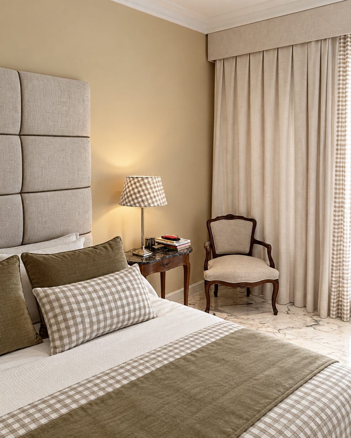 Dormitorio con cabecero tapizado a medida en paneles de tejido beige y butaca clásica junto a la ventana